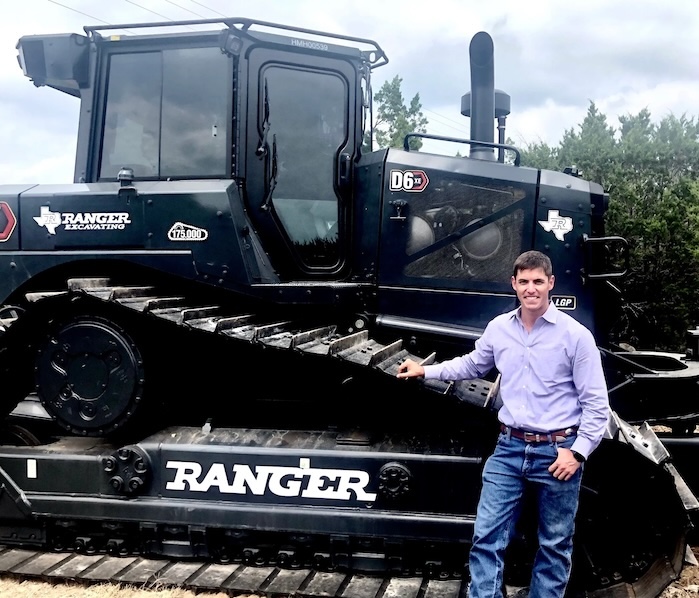 Will Romo dressed in blue jeans and a button down, standing next to a black Ranger Excavating digger.